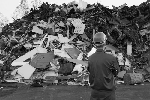 A man wearing a safety helmet and dark work clothing stands in front of a large pile of mixed construction and household waste on an outdoor paved surface. The waste includes broken and discarded white panels, plastic items, metal scraps, and other debris, forming a densely stacked mound extending into the background. The scene appears to be part of a rubbish clearance operation, reflective of services offered by Office Clearance Notting Hill in the local area near the postcode NW10 within London. The overall setting is outdoor, with natural lighting illuminating the scene, emphasizing the variety of materials and textures in the waste pile, which includes both smooth and jagged surfaces. The man’s position in the foreground provides a clear perspective of the scale of the waste accumulation, indicating a significant clearance task. This image highlights the importance of professional rubbish removal services for managing bulky or hazardous waste in the local vicinity.
