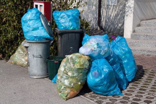 A collection of various rubbish bags and bins positioned on a paved driveway in an outdoor urban setting, with some blue plastic bags filled with waste, and other smaller black, grey, and green containers. The blue bags contain mixed waste, possibly household or office rubbish, and are stacked or leaning against the bins. The grey bins are made of metal or plastic, with one having a closed lid. The environment includes a background of bushes, a grey wall, and a set of concrete steps leading upward. The scene is illuminated by natural daylight, highlighting the different textures of the bags and bins, and indicative of waste disposal or rubbish removal activity typical of local services in Notting Hill, London. This visual aligns with the services offered by Office Clearance Notting Hill, specialising in rubbish collection and clearance for commercial or residential properties in the area.