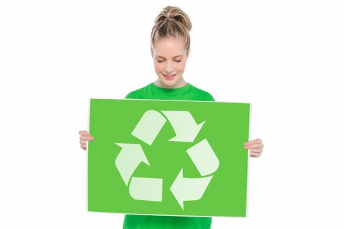 A young woman with blonde hair tied back, wearing a bright green shirt, is smiling while holding a large rectangular sign featuring the universal recycling symbol composed of three white arrows forming a triangle on a vibrant green background. The background of the image is plain white, and she appears to be promoting environmental awareness or waste recycling. The image emphasizes the importance of recycling materials as part of rubbish removal and environmental conservation efforts, similar to services offered by companies like Office Clearance Notting Hill, which operate within the W11 postal district of London. Her cheerful expression and the clear recycling symbol underscore a positive message for waste management and sustainable disposal practices, aligning with the principles of responsible rubbish clearance and environmental care consistent with local waste services in Notting Hill, London.