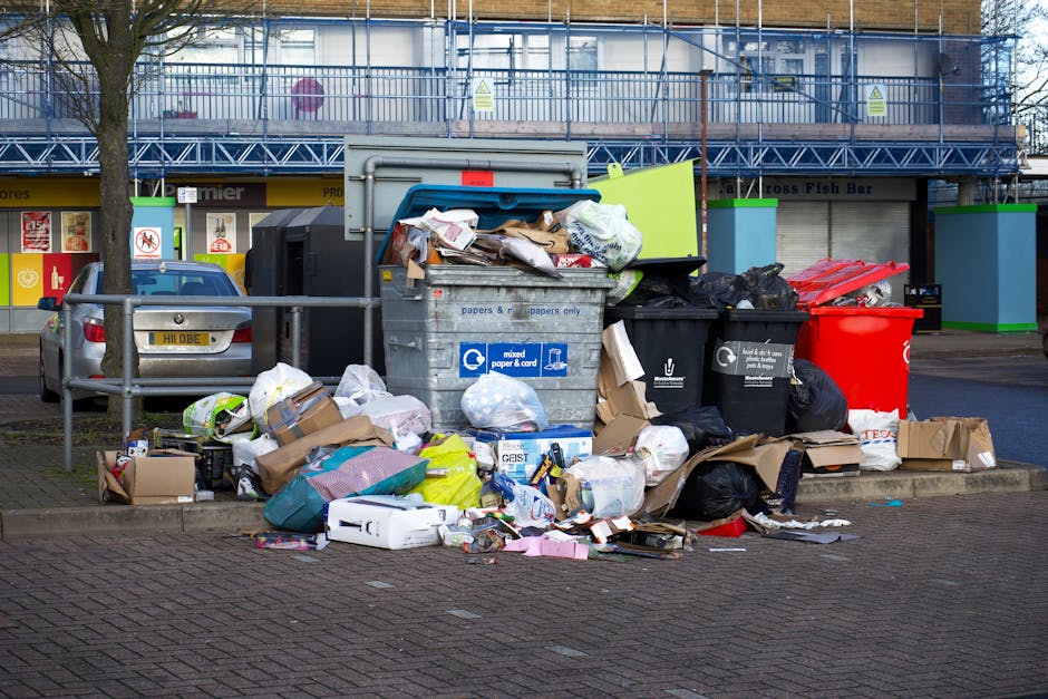 A collection of black rubbish bags filled with waste and discarded packaging materials stacked in front of a red metal door on an exterior wall. The bags are piled on the pavement, with some leaning a