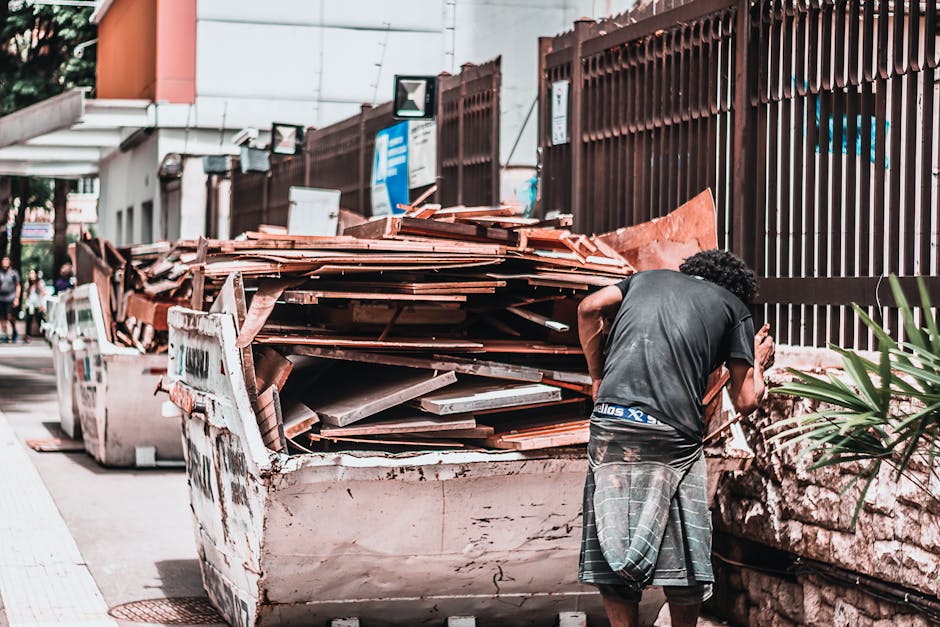 A sanitation worker wearing an orange and yellow reflective safety vest and dark trousers operates a red refuse collection vehicle parked on the side of a street, with the vehicle's rear hatch open re