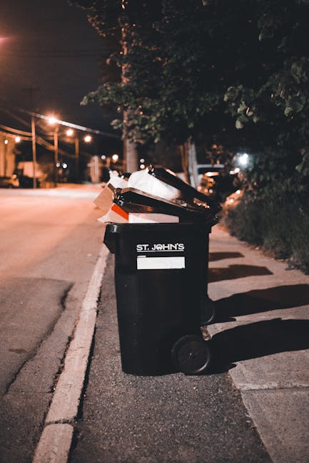 A large pile of mixed household waste and rubbish is accumulated in front of a commercial building, consisting of white, black, and red bin liners, cardboard boxes, paper, and plastic packaging, some